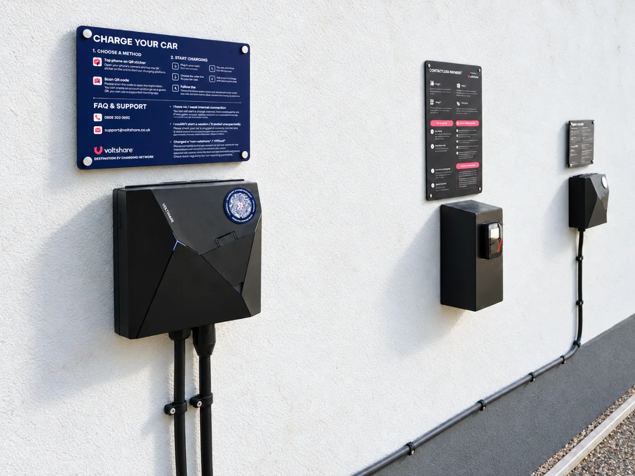 A black voltshare wall charger under a navy sign headed Charge your car, with the contactless payment terminal and another charger further along the wall