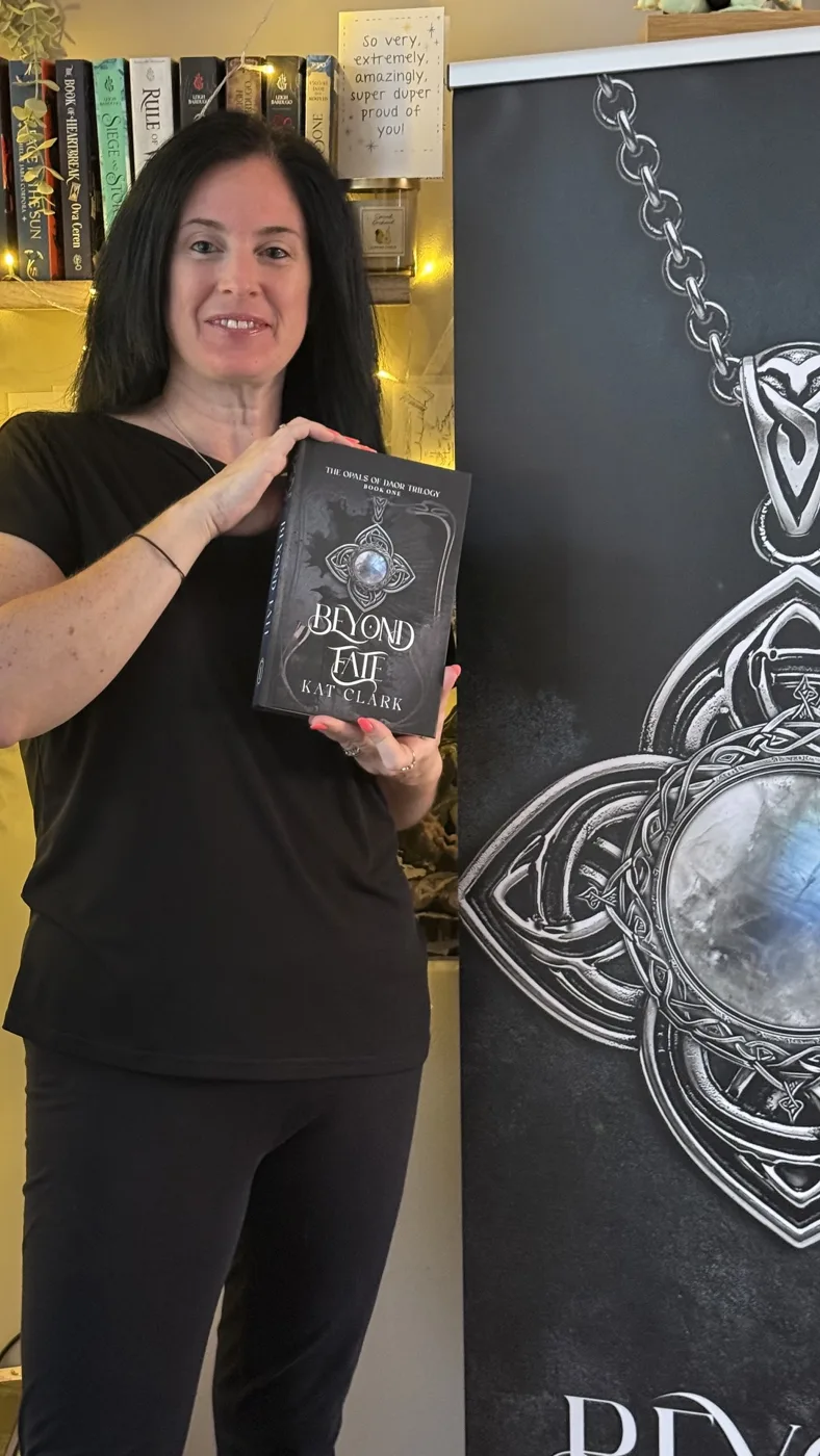 Kat Clark holding a hardback copy of Beyond Fate beside a banner showing the book's Celtic pendant