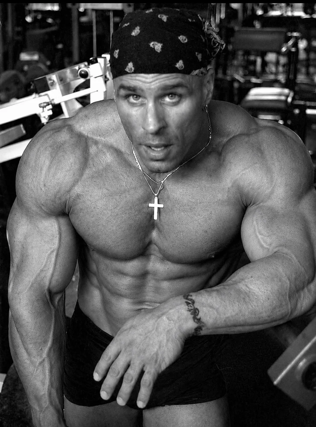 Black and white photograph of John Xuereb, a muscular bodybuilder in a bandana and cross necklace, leaning forward in a gym